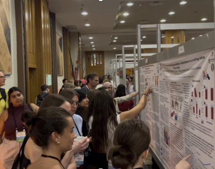 Conference attendees gather in a crowded poster session, reading and discussing student research displays. Several participants point to charts and text on the posters while others wait to speak with presenters.